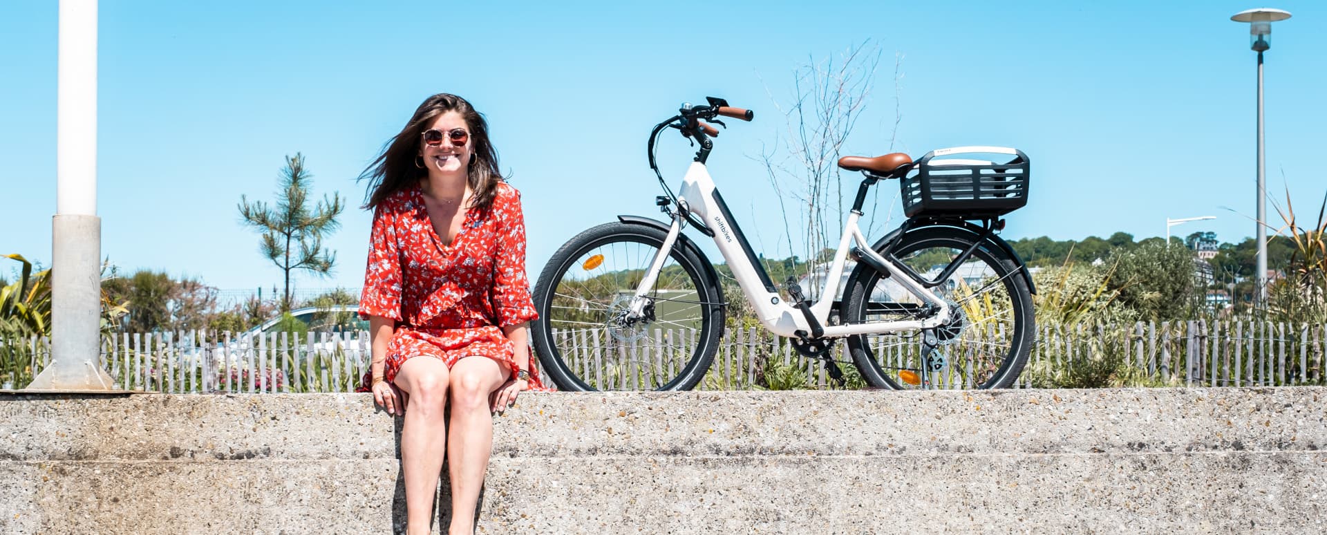 une femme en robe rouge avec des lunettes de soleil est assise avec son vélo électrique derrière elle